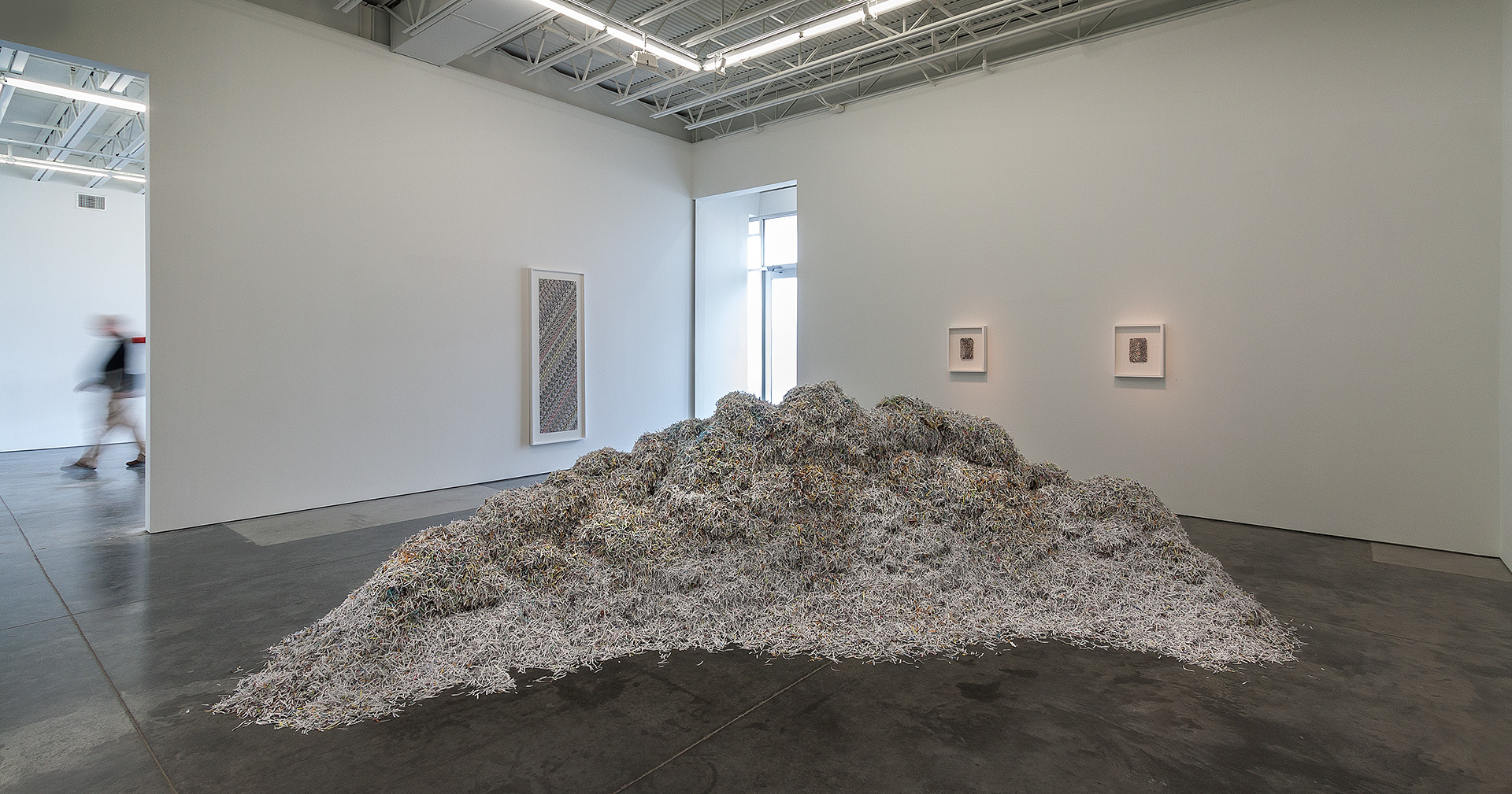 A large mound of shredded gray and white paper material dominates the center of a white gallery space, with framed works on the surrounding walls and a visitor visible in the background.