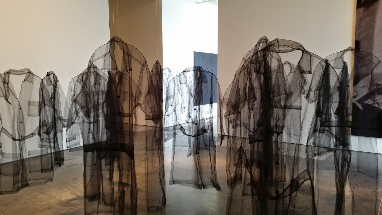 Translucent dark fabric forms layered silhouettes of human figures displayed in a gallery space, creating ghostly overlapping profiles against white walls and illuminated by natural light from above.