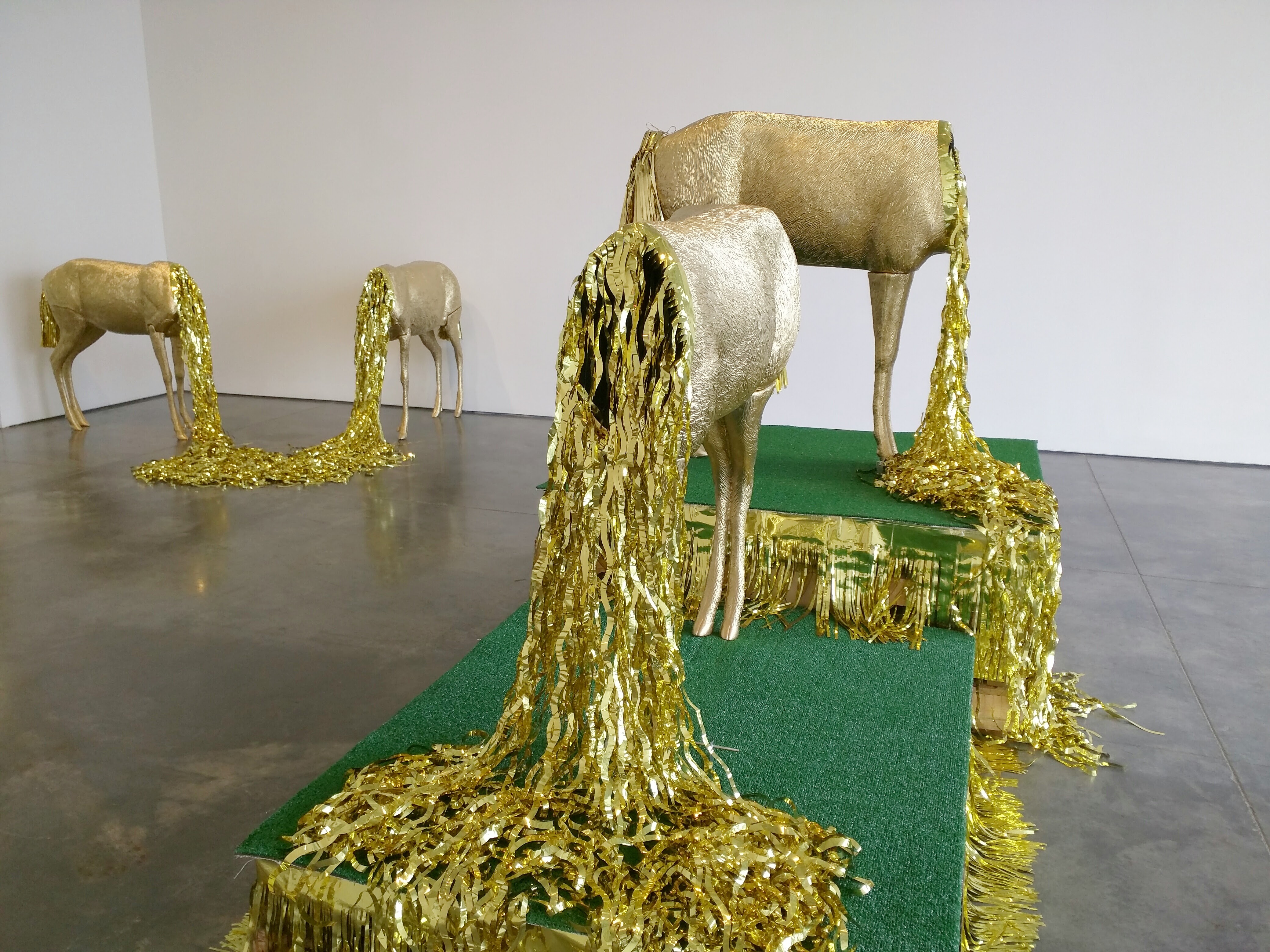 Four gilded deer sculptures of varying sizes stand on green platforms, their bodies cast in metallic gold with dramatic strands of golden fringe cascading from their necks and bodies onto the gallery floor, creating an ornate, ceremonial appearance against white walls and polished concrete.