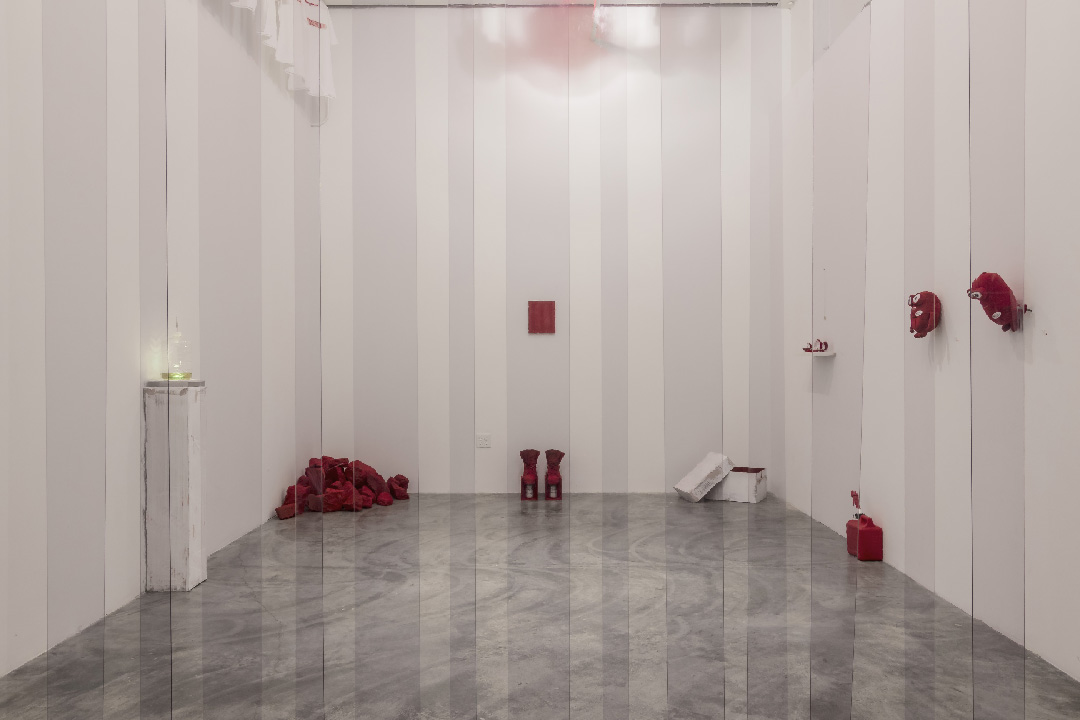 Minimalist gallery installation featuring scattered crimson objects and sculptural forms arranged across a polished concrete floor against white walls with vertical paneling. A single glass vessel with green plant material stands isolated on the left, while red geometric shapes, fabric bundles, and vessel-like forms are distributed throughout the sparse, contemplative space.