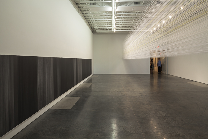 A minimalist gallery installation featuring a dense black linear drawing mounted along the lower left wall and a delicate cloud of fine white threads suspended from the ceiling across the gallery's length, creating an immersive environmental work that activates the full white-walled space with industrial ceiling visible above.