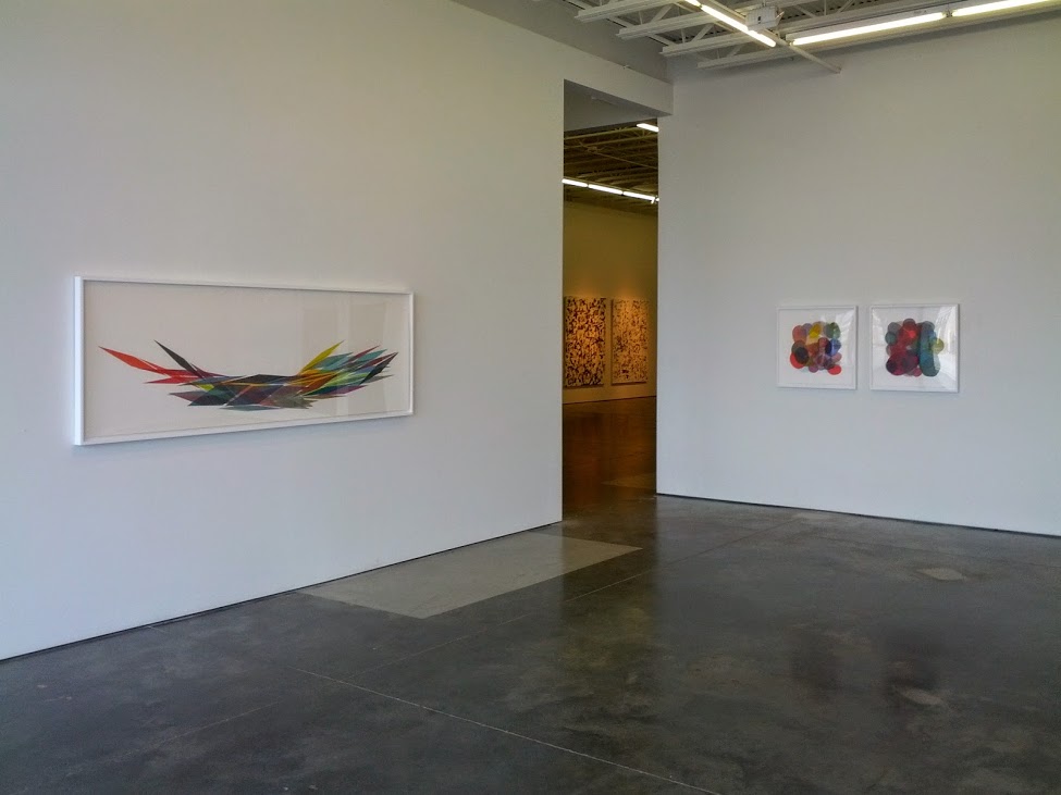 A minimalist gallery interior displays abstract artworks on white walls. On the left, a horizontal framed piece features intersecting lines in red, yellow, black, and blue radiating outward. On the right wall, two smaller framed prints show organic clusters of colorful circles in jewel tones. A doorway with warm lighting opens to an adjacent gallery space beyond.