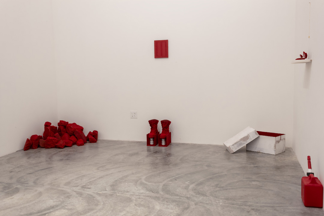 A minimalist gallery installation featuring scattered red fabric objects and white boxes arranged across a polished concrete floor. Red elements include crumpled cloth on the left, two upright boot-shaped forms in the center, a red canister on the right, and a small red rectangle mounted on the upper wall, contrasted by white storage boxes and neutral gallery walls.