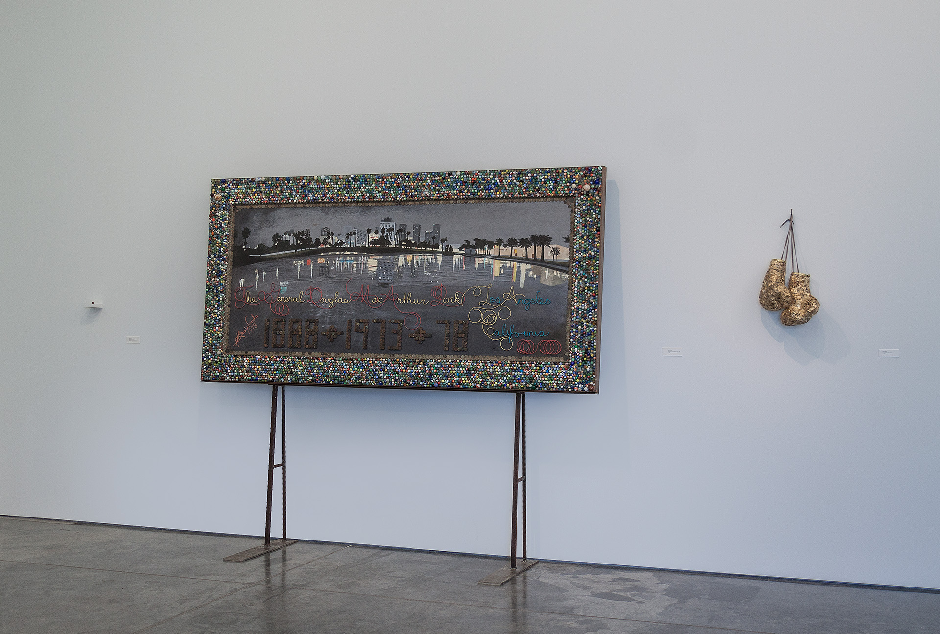 Mixed-media artwork on a stand featuring a photograph of a lakeside cityscape with a beaded, multicolored border frame. The lower portion contains embossed text and decorative elements in gold and red. To the right hangs a separate sculptural piece made of gathered, gilded fabric suspended by thin wire.