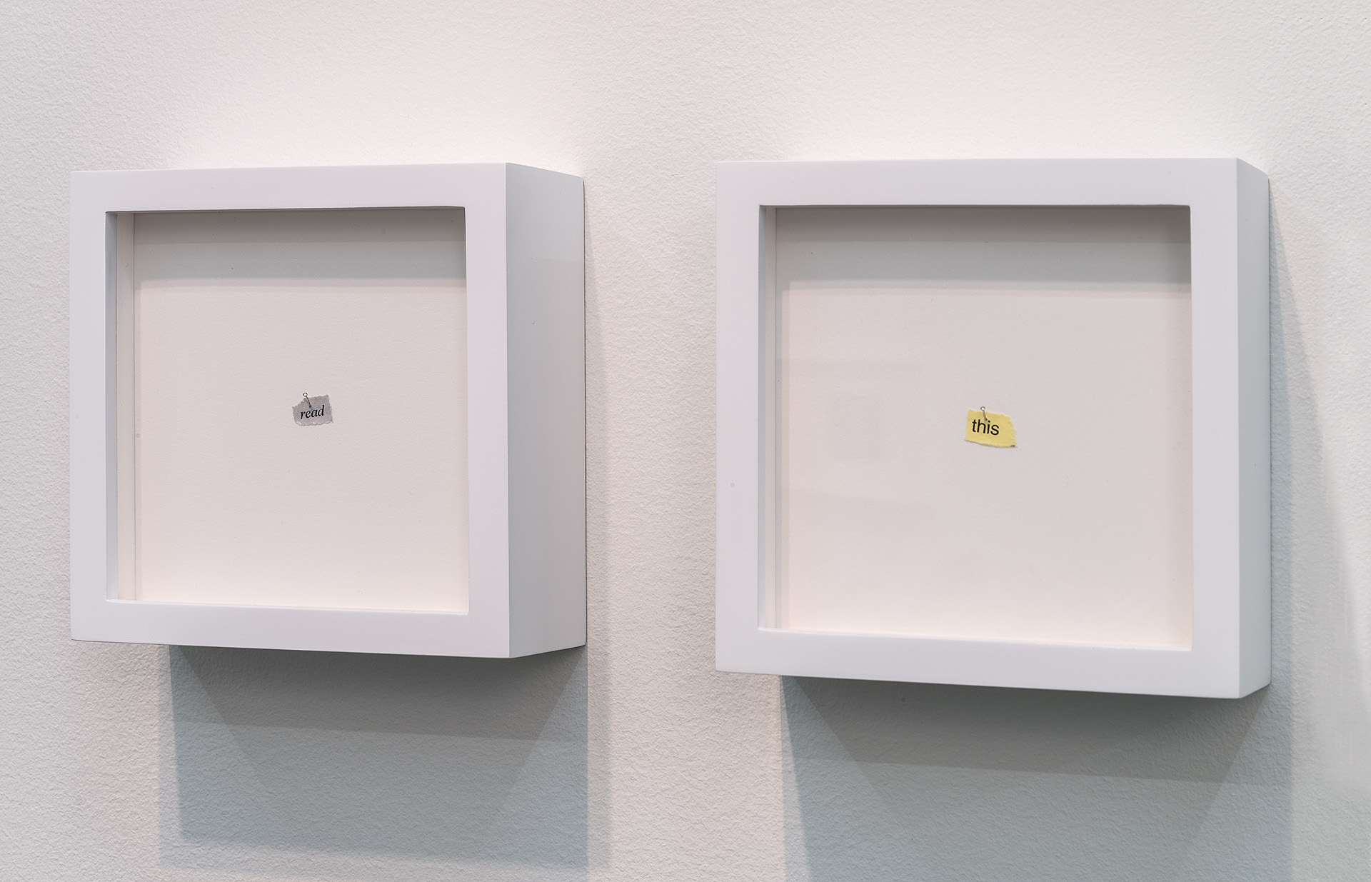 Two white box-shaped wall-mounted structures with recessed glass fronts containing cream-colored backing, each displaying a small torn piece of printed text—one reading "read" in gray lettering, the other "this" in yellow—positioned side by side against a white wall.